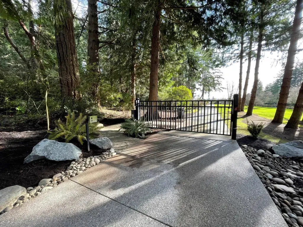 Automated metal swing gate across a residential driveway lined with evergreen trees and river-rock borders